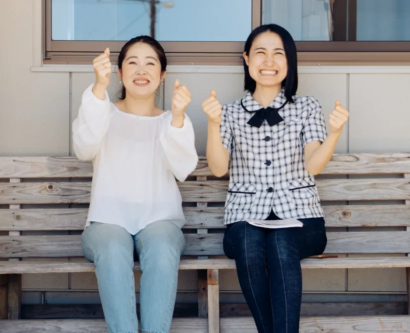 教習所内の長椅子に並んで座って、ハンドルを握っているポーズをしている女性教習生と女性指導員