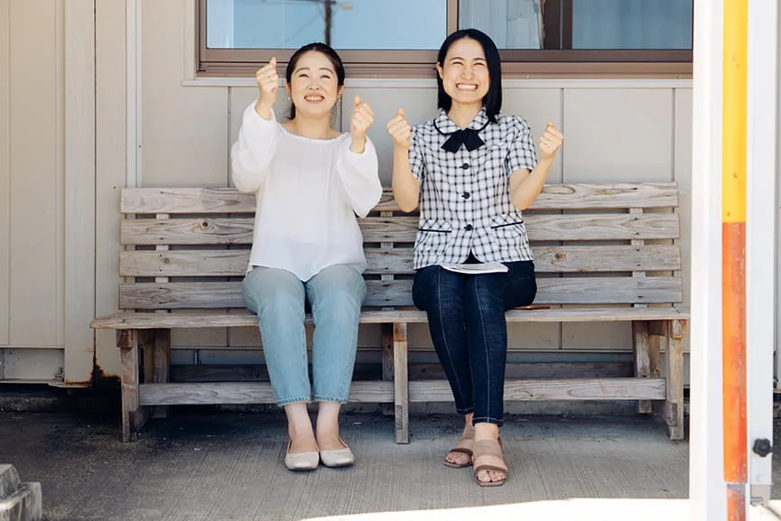 構内の長椅子に座って、ハンドルを握るポーズをとる女性教習生と女性指導員