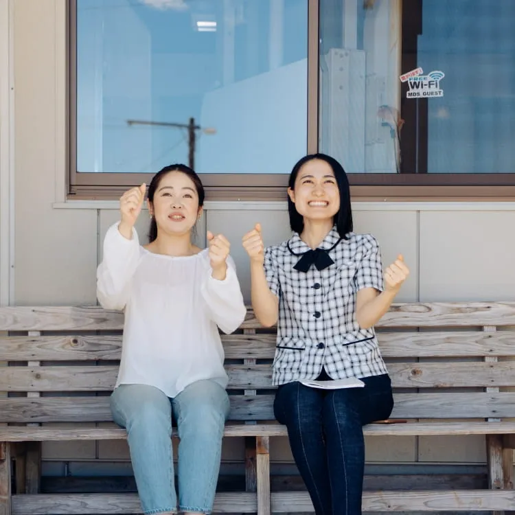 構内の長椅子に座って、ハンドルを握るポーズをとる女性教習生と女性指導員