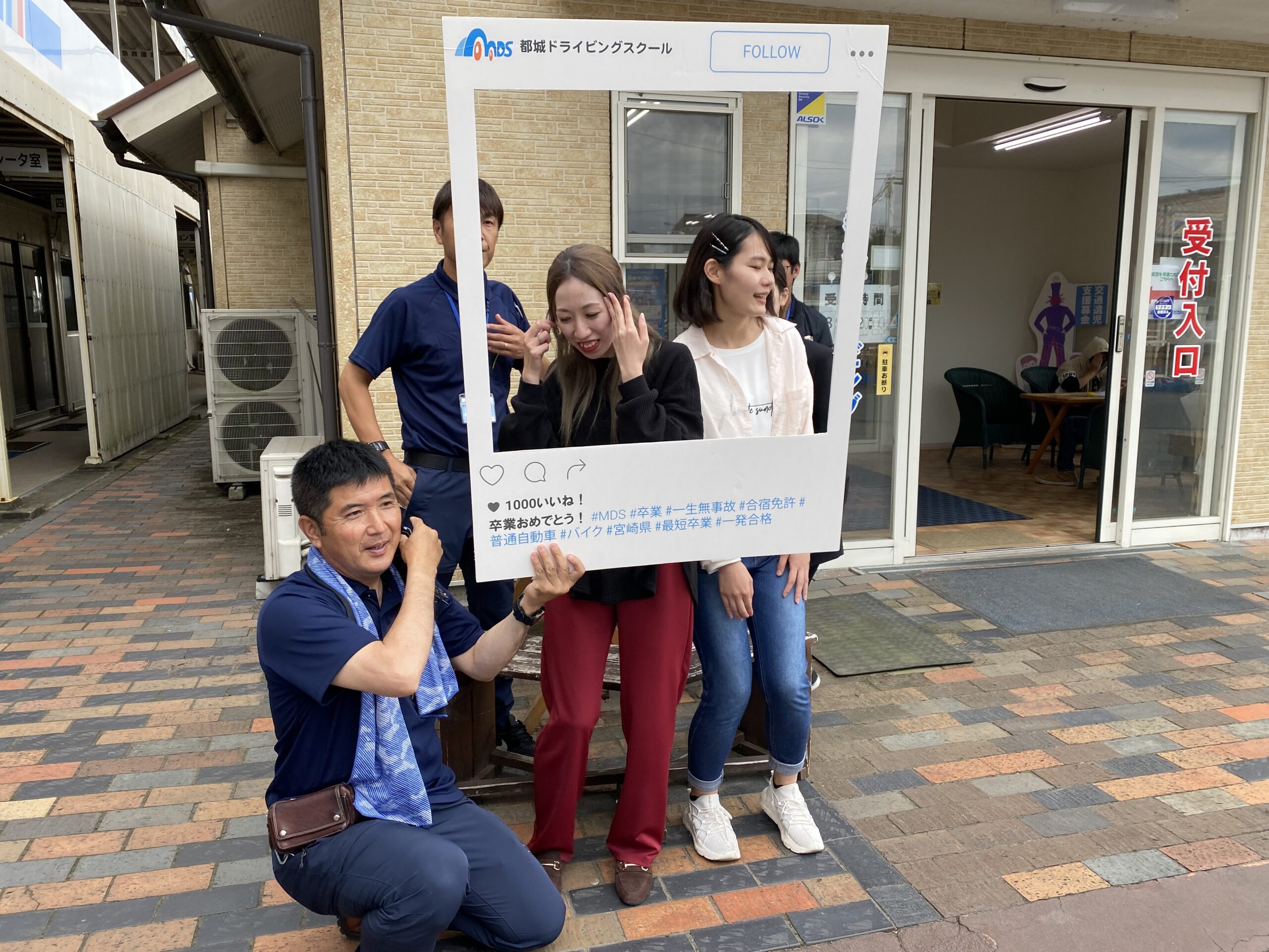 都城ドライビングスクールの受付入り口前で指導員がインスタの枠を持って、教習生と和やかに写真を撮っている様子